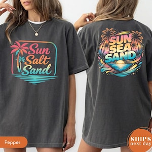May include: A charcoal gray t-shirt featuring a graphic on the front and back. The front graphic shows a palm tree and the text Sun Salt Sand in a colorful gradient. The back graphic displays a sunset, palm trees, and the text Sun Sea Sand.