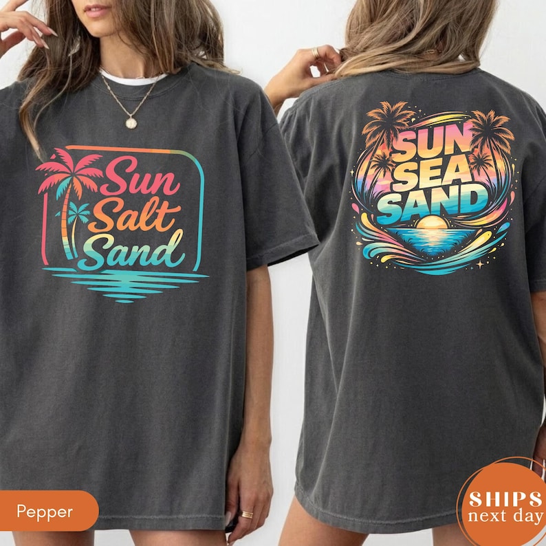 May include: A charcoal gray t-shirt featuring a graphic on the front and back. The front graphic shows a palm tree and the text Sun Salt Sand in a colorful gradient. The back graphic displays a sunset, palm trees, and the text Sun Sea Sand.
