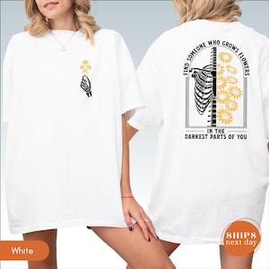 A white t-shirt with a graphic design on the back, depicting a skeleton with flowers and the text "Find someone who grows flowers in the darkest parts of you".