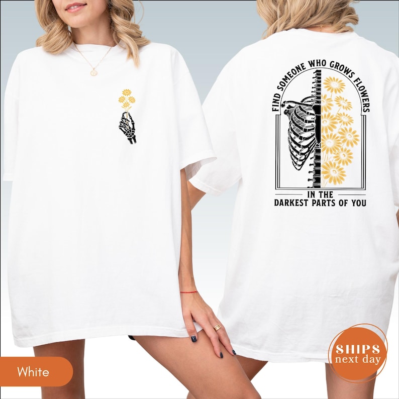 A white t-shirt with a graphic design on the back, depicting a skeleton with flowers and the text "Find someone who grows flowers in the darkest parts of you".