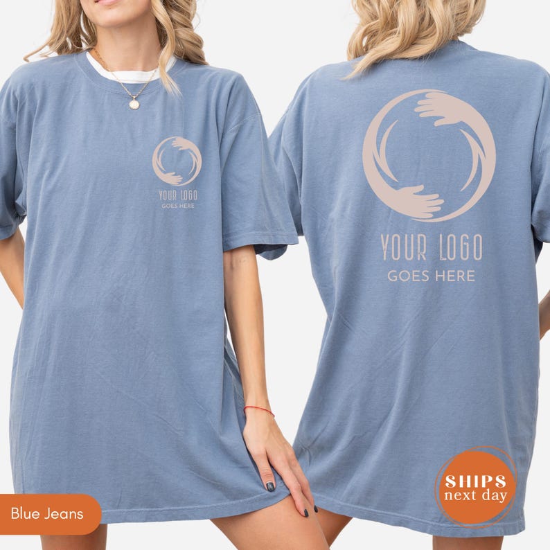 May include: A dusty blue short-sleeve t-shirt featuring a circular graphic of two hands forming a ring. The front left chest displays a small version of the logo, while the back features a large version with the text YOUR LOGO GOES HERE.