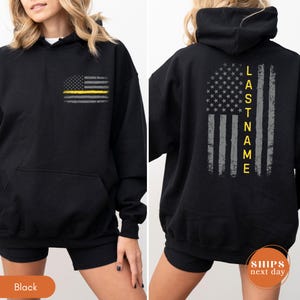 May include: A black hooded sweatshirt featuring a distressed American flag graphic on the front left chest with a yellow stripe. The back displays a large distressed American flag graphic with a yellow stripe and the vertical text LASTNAME.