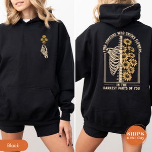 May include: Black hoodie with a front pocket and a hood. The front features a skeleton hand holding yellow flowers. The back displays a rib cage with yellow flowers and the text: "FIND SOMEONE WHO GROWS FLOWERS IN THE DARKEST PARTS OF YOU."