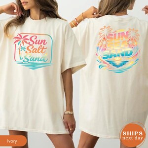 May include: An ivory t-shirt featuring a front graphic with a palm tree and the text Sun Salt Sand in a gradient of pink, orange, and blue. The back graphic displays a larger, circular beach scene with palm trees and the text Sun Sea Sand.