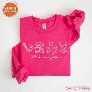 May include: A bright pink long-sleeve sweatshirt featuring a white graphic print of baking tools including a whisk, measuring spoons, a stand mixer, an apron, a rolling pin, and a piping bag, with the text "I'll be in my office" printed below.