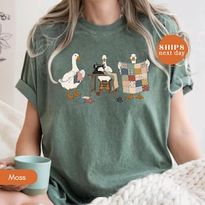 May include: A moss green t-shirt featuring a graphic of three white geese engaged in craft activities. One goose knits, another operates a sewing machine, and the third holds up a colorful patchwork quilt. The design includes small fabric scraps.