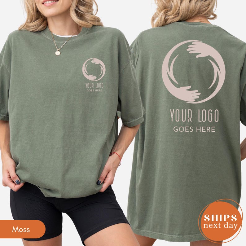 May include: A moss green t-shirt featuring a small circular logo of two hands forming a circle on the left chest and a larger version of the same logo on the back. Below both logos, the text reads YOUR LOGO GOES HERE in a sans-serif font.