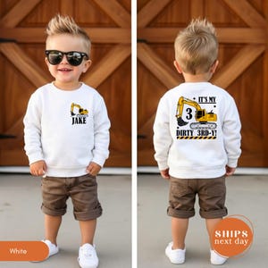 May include: A white long-sleeve sweatshirt featuring a yellow excavator graphic on the front left chest with the name Jake printed below. The back displays a large yellow excavator graphic with the text It's My 3 Dirty 3rd-Y!