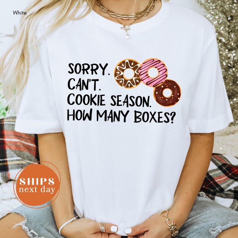 A person wearing a white t-shirt with a humorous text printed on it, which reads "Sorry, can't. Cookie season. How many boxes?" The person is also wearing a necklace and has a bracelet on their wrist.