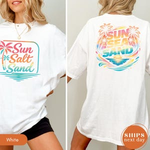 May include: A white t-shirt featuring a colorful graphic on the front and back. The front design shows palm trees and the text Sun Salt Sand in a gradient. The back design displays a larger circular graphic with palm trees and the text Sun Sea Sand.
