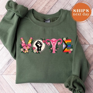 May include: An olive green crewneck sweatshirt featuring the word VOTE in a graphic design. The V is formed by two pencils with a pink bow, the O is a black raised fist, the T is a pink uterus illustration, and the E is a rainbow pride flag.