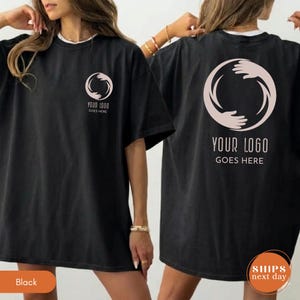 May include: A black t-shirt featuring a light pink circular graphic of two hands reaching toward each other. The front chest displays a small version of this logo with the text YOUR LOGO GOES HERE, while the back features a large version of the same logo.