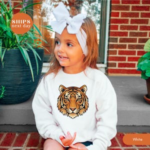 May include: A white sweatshirt featuring a detailed tiger face graphic in shades of brown and orange. The sweatshirt is worn by a child with a white bow headband. The text "Ships next day" is visible in the upper left corner, and the word "White" is in the lower right corner.