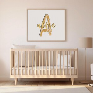 May include: A framed print featuring the name "alfie" in a stylized font. The letter "A" is filled with a tan and cream zebra stripe pattern. The name is in a gold script with a dark blue shadow. A light wood crib is in the foreground.