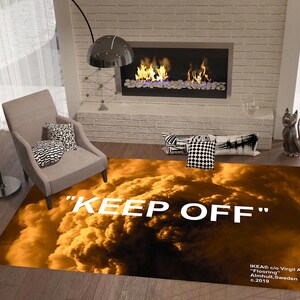 以下が含まれることがあります： オレンジと茶色の雲模様と白い文字「KEEP OFF」が特徴の長方形のラグ。暖炉、椅子、フロアランプのある部屋に敷かれています。ラグには「IKEA c/o Virgil Abloh」と表示されています。