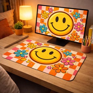 Op de afbeelding: Een bureau-opstelling in retrostijl met een computermonitor en een bijpassende muismat. De monitor en muismat tonen een grote gele smiley omringd door kleurrijke bloemen en sterren op een oranje geruite achtergrond.