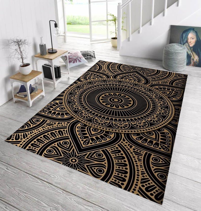 May include: A rectangular area rug with a black and tan mandala design. The rug features a central circular pattern with intricate details and a repeating floral motif. The rug is placed on a light-colored wooden floor.