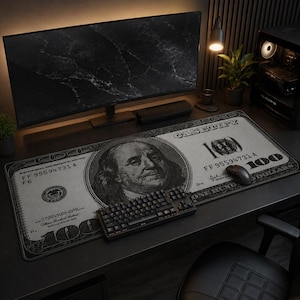May include: A black and white desk mat featuring a design of a $100 bill, with a portrait of Benjamin Franklin. A black keyboard and mouse are on the mat. A computer monitor and other desk accessories are in the background.
