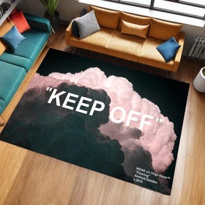 Op de afbeelding: Een rechthoekig vloerkleed met een wolkenontwerp in roze, wit en grijs, met de woorden "KEEP OFF" in grote witte letters. Het vloerkleed ligt op een houten vloer. De tekst "IKEA c/o Virgil Abloh" is zichtbaar.