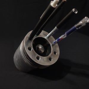 Könnte beinhalten: Ein silberner Stifthalter aus Metall in Form eines Motorzylinders mit vier Löchern für Stifte. Drei Stifte befinden sich im Stifthalter, ein schwarzer, ein weißer und ein blauer.