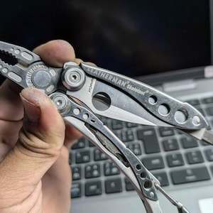 May include: A silver Leatherman Skeletool multi-tool with pliers, a knife, and other tools. The multi-tool is partially open, revealing the knife blade and other components. The brand name is visible on the tool.