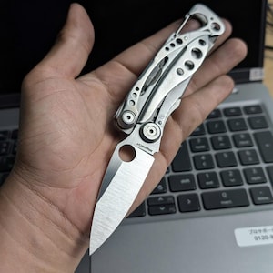 May include: A silver folding knife with a partially open blade, held in a hand. The knife has a textured handle and a sharp, pointed blade. The blade is approximately 3 inches long. The knife is resting on a laptop keyboard.