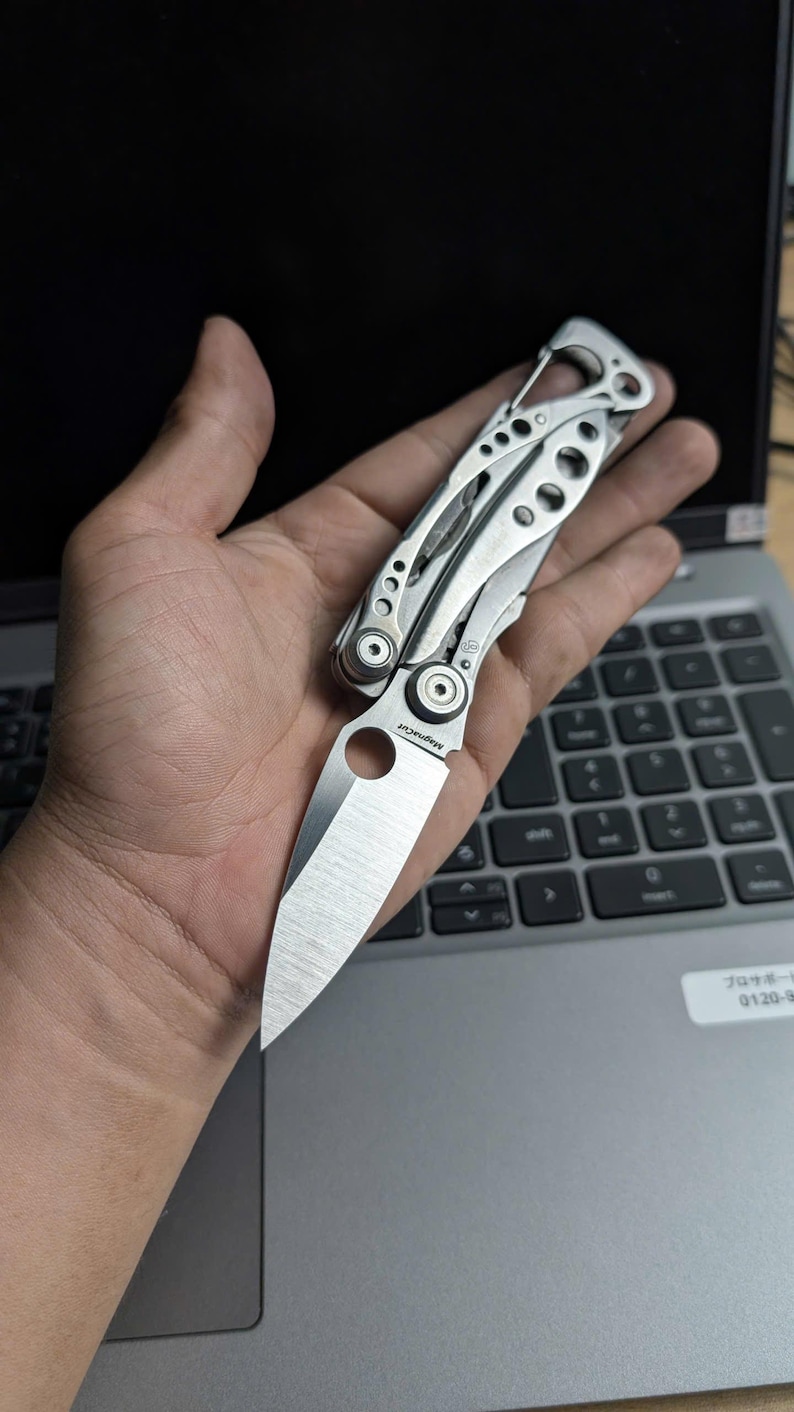 May include: A silver folding knife with a partially open blade, held in a hand. The knife has a textured handle and a sharp, pointed blade. The blade is approximately 3 inches long. The knife is resting on a laptop keyboard.