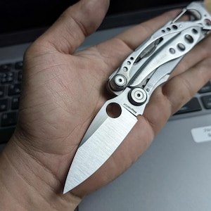 Może przedstawiać: Srebrny nóż wielofunkcyjny z częściowo otwartym ostrzem, trzymany w dłoni. Nóż posiada różnorodne narzędzia i teksturowany uchwyt. Ostrze jest oznaczone "Magnacut". Nóż spoczywa na laptopie.