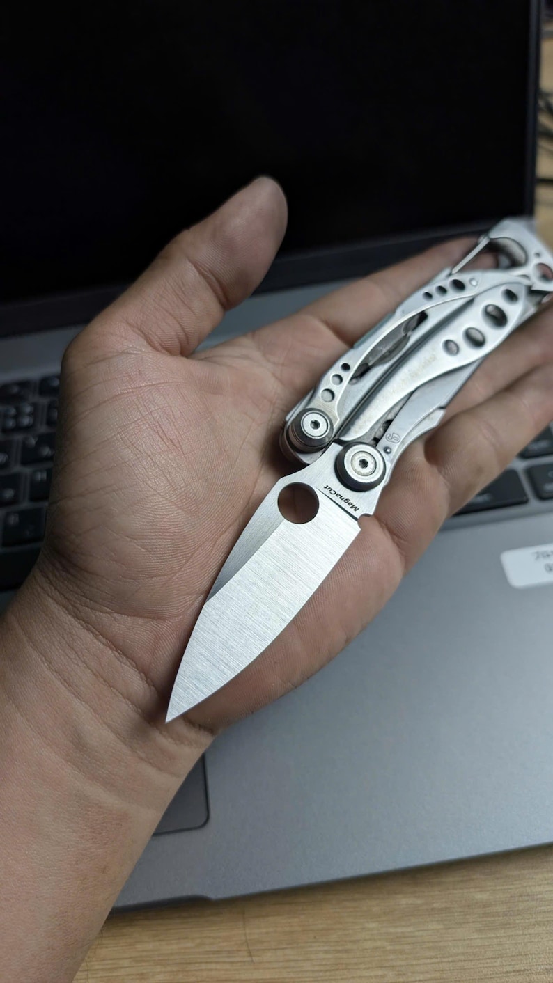May include: A silver multi-tool knife with a partially open blade, held in a hand. The knife has a variety of tools and a textured handle. The blade is marked "Magnacut". The knife is resting on a laptop.