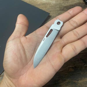 May include: A silver knife blade with a hole in the handle. The blade is about 3 inches long and has a slight curve. The blade is marked with the text "90V".