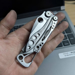 May include: A silver multi-tool with a variety of implements, including a knife blade, is held open in a hand. The tool has a carabiner clip and several circular cutouts. The tool is resting on a laptop.