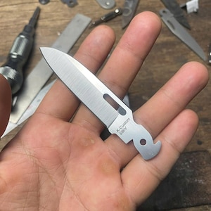May include: A small, silver, pocket knife with a blade and a hook on the end. The knife has the text "K-Custom S90V" engraved on the side.