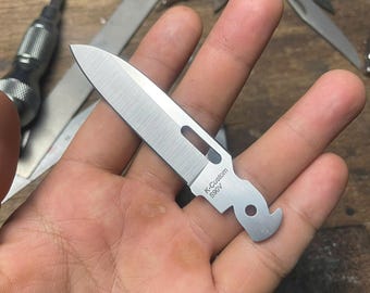 May include: A small, silver, pocket knife with a blade and a hook on the end. The knife has the text "K-Custom S90V" engraved on the side.