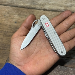 Op de afbeelding: Een zilverkleurig multifunctioneel zakmes met een gestructureerd Alox-handvat voorzien van een rood-wit kruis-embleem. Het mes heeft één gedeeltelijk uitgeklapt roestvrijstalen lemmet met een duimgat en de MagnaCut-staalmarkering is zichtbaar.