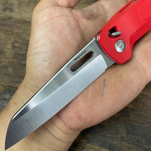 以下が含まれることがあります： 銀色の刃が付いた赤い折りたたみナイフ。刃には「MagnaCut」の文字が印刷されています。ナイフは開いており、鋭い刃先が見えています。ハンドルは鮮やかな赤色です。
