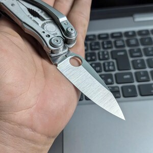 May include: A silver multi-tool with a partially open blade. The blade has a hole near the base. The tool is held in a hand, with a laptop keyboard in the background. The tool appears to be made of stainless steel.