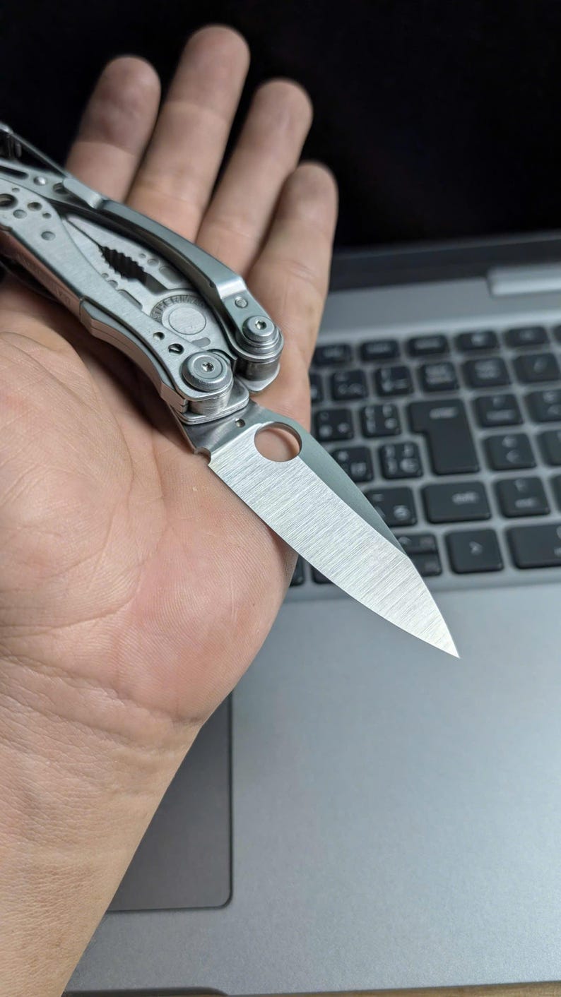 May include: A silver multi-tool with a partially open blade. The blade has a hole near the base. The tool is held in a hand, with a laptop keyboard in the background. The tool appears to be made of stainless steel.