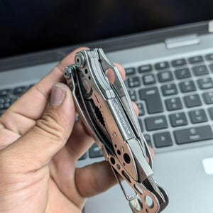 May include: A multi-tool with a silver finish, featuring pliers, a knife, and other tools. The brand name "LEATHERMAN" is visible on the side. The tool is held against a laptop keyboard.