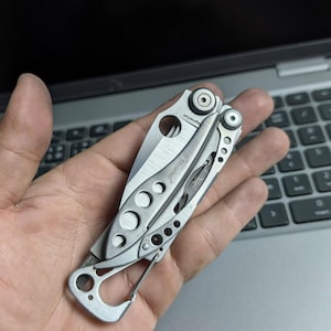 May include: A silver multi-tool with a partially open blade and a carabiner clip. The tool has a series of circular cutouts and the text "Leatherman Skeletool" is visible. The tool is designed for various tasks.