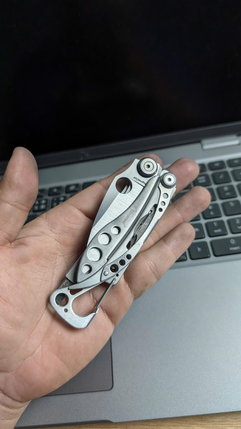 May include: A silver multi-tool with a partially open blade and a carabiner clip. The tool has a series of circular cutouts and the text "Leatherman Skeletool" is visible. The tool is designed for various tasks.