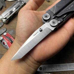 May include: A black multi-tool with a silver blade that says "Megacut" on it. The tool is open and the blade is extended. The tool is being held in a person's hand.