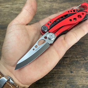 Könnte beinhalten: Ein rotes Multitool mit schwarzem Griff und einer silbernen Klinge. Die Klinge ist teilweise ausgefahren und trägt die Aufschrift "MagniCut".