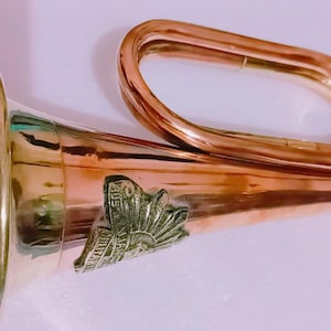 May include: A copper-colored bugle with a flared bell and a curved mouthpiece. The bugle features a decorative emblem near the bell and a small mouthpiece with a chain. The instrument is designed for making musical sounds.