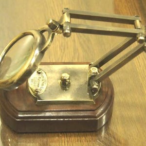 May include: A brass magnifying glass with a wooden base and a hinged arm. The magnifying glass is labelled "Harris & Son Ltd. Princess St. London, E.C.1."
