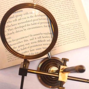 May include: A brass magnifying glass with a stand, focused on a book page with the text "mind group with can you who will aid you in the development who have accumulated great fortunes V. They developed the habit of persistence closely driven by circumstances, that the".