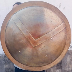 May include: A round, bronze-coloured shield with a distinctive V-shaped design in the centre. The shield has a raised rim and a textured surface.