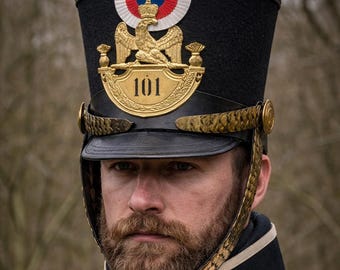 Puede incluir: Un sombrero militar negro con un pompón rojo, un emblema de águila dorada y el número 101. El sombrero forma parte de un uniforme rojo con correas blancas y detalles dorados. La persona tiene barba.