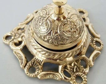 May include: A gold-toned metal service bell with an ornate base and a detailed floral design. The bell has a round top with a knob on top.