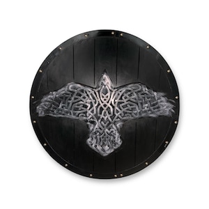 May include: A circular black shield featuring a central silver-toned raven emblem with intricate knotwork patterns. The shield surface displays vertical plank lines and is bordered by a series of small, evenly spaced metallic studs along the edge.