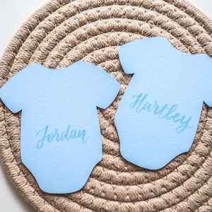 May include: Two light blue paper cutouts shaped like baby bodysuits. Each cutout features a name printed in a darker blue script font. One cutout displays the name Jordan, and the other cutout displays the name Hartley, both centered on the front.